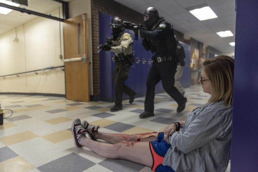 A woman sitting on the floor of a hallway pretending to be dead while police sweep the corridors of a school during a drill. 